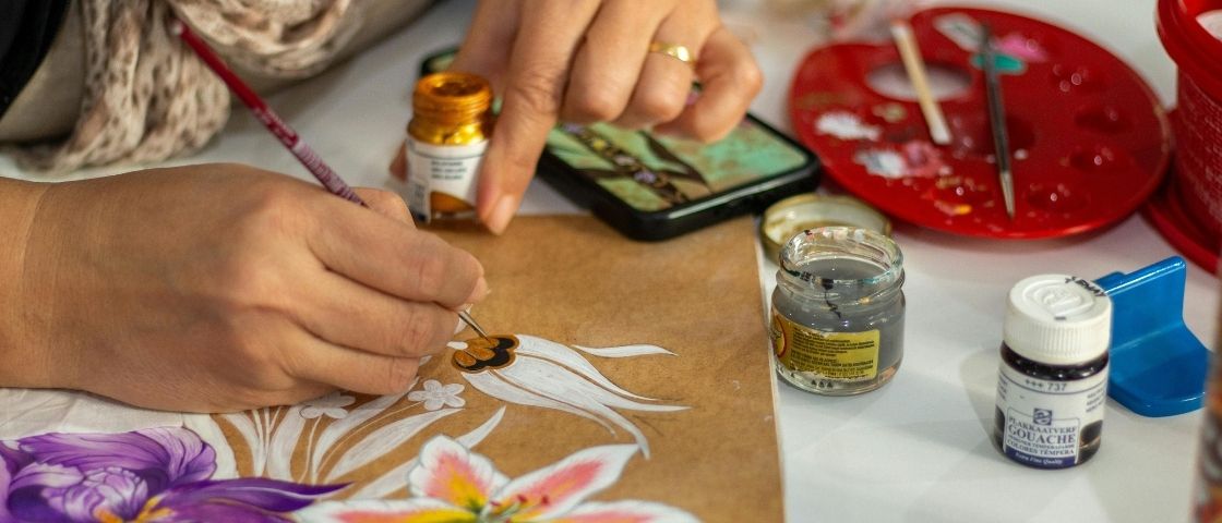 Main peignant des fleurs colorées à la gouache sur papier kraft, avec pots de peinture autour.