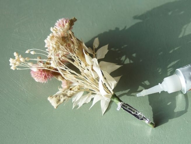 Boutonnière en fleurs séchées terminée avec une attache métallique, à côté d’un tube de colle.