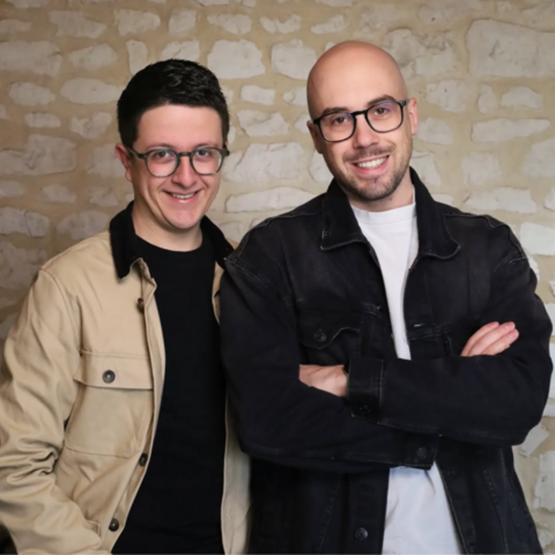 Deux hommes souriants portant des lunettes posent devant un mur en pierre, bras croisés.