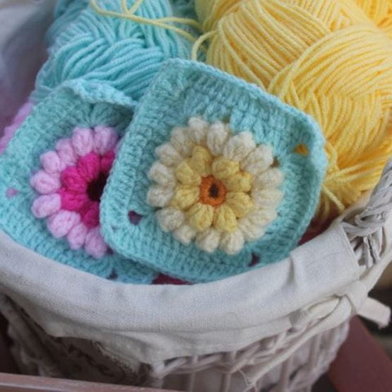 Gros plan sur un panier contenant des pelotes de laine jaune et bleue avec deux granny squares crochetés aux motifs floraux.