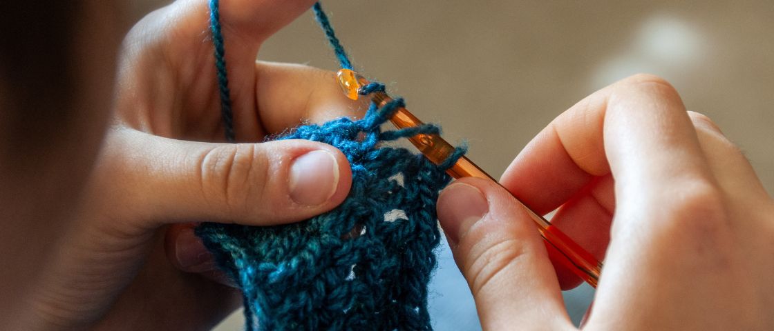 Mains en train de crocheter une pièce en laine bleue avec un crochet orange.